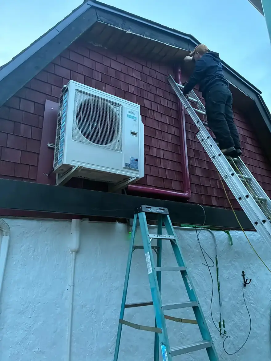 Technician on a ladder beside a wall-mounted heat pump outdoor unit